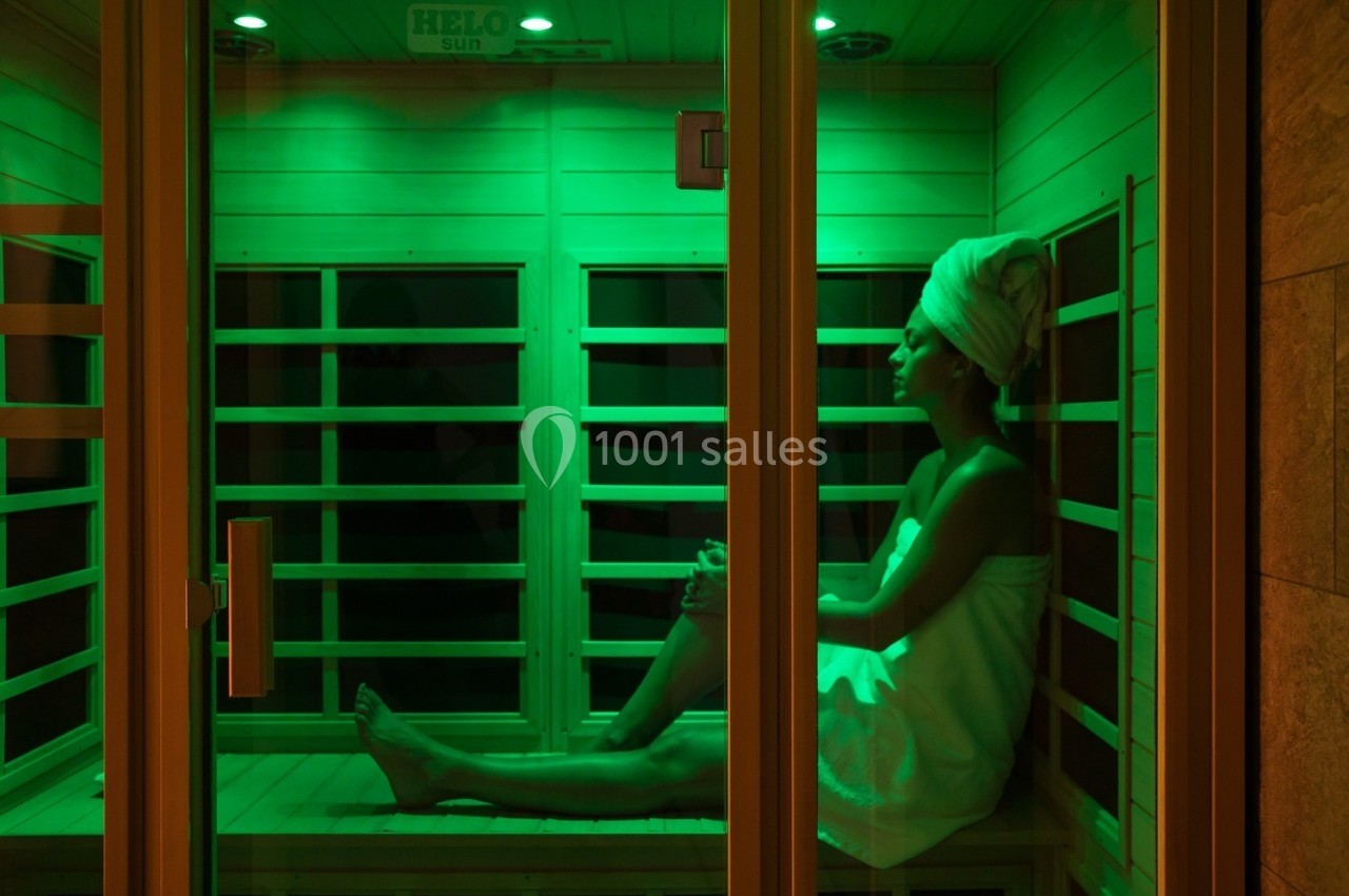 Femme assise dans un sauna à lumière verte, enveloppée dans une serviette et portant une serviette sur la tête.