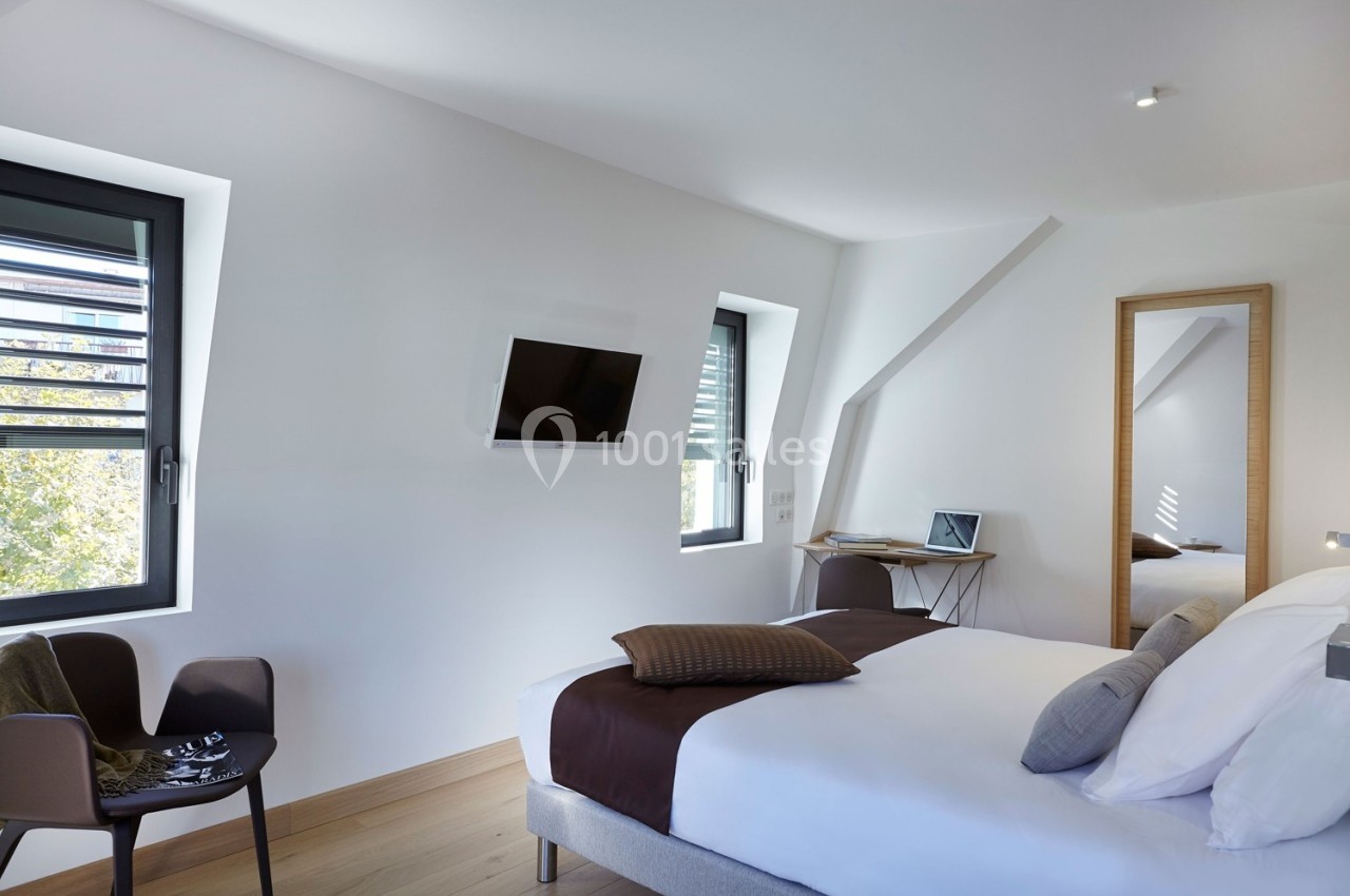 Chambre lumineuse avec lit double, bureau, chaise, télévision murale et grandes fenêtres laissant entrer la lumière…