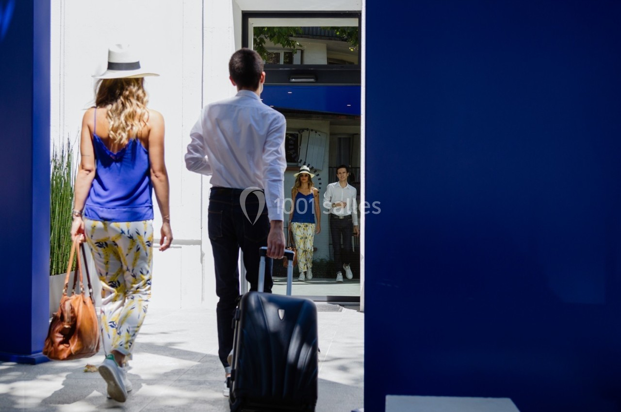 Un homme et une femme entrent dans un bâtiment moderne, lui portant une valise, elle un sac à main.
