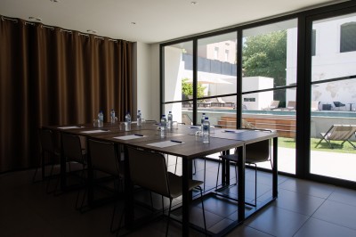 Chambre lumineuse avec lit double, table en bois, chaise beige, miroir et grande fenêtre avec stores.