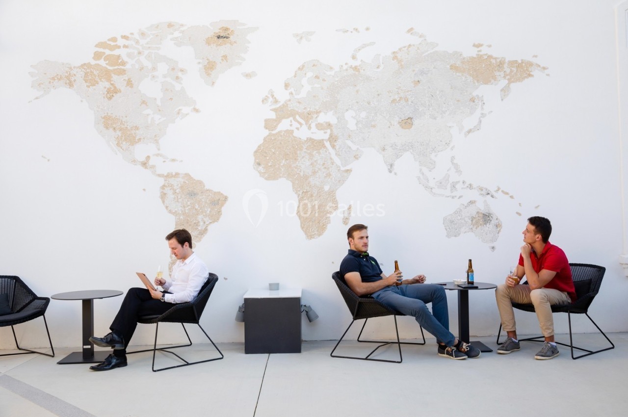 Trois hommes assis sur des chaises devant une carte du monde murale, discutant ou utilisant un ordinateur portable.