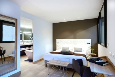 Chambre lumineuse avec lit double, table en bois, chaise beige, miroir et grande fenêtre avec stores.