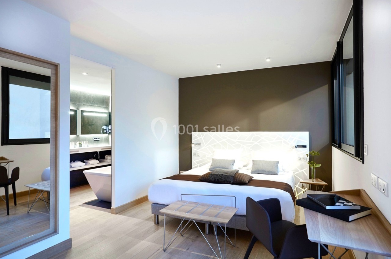 Chambre moderne avec lit double, bureau, miroir, et salle de bain attenante équipée d'une baignoire.