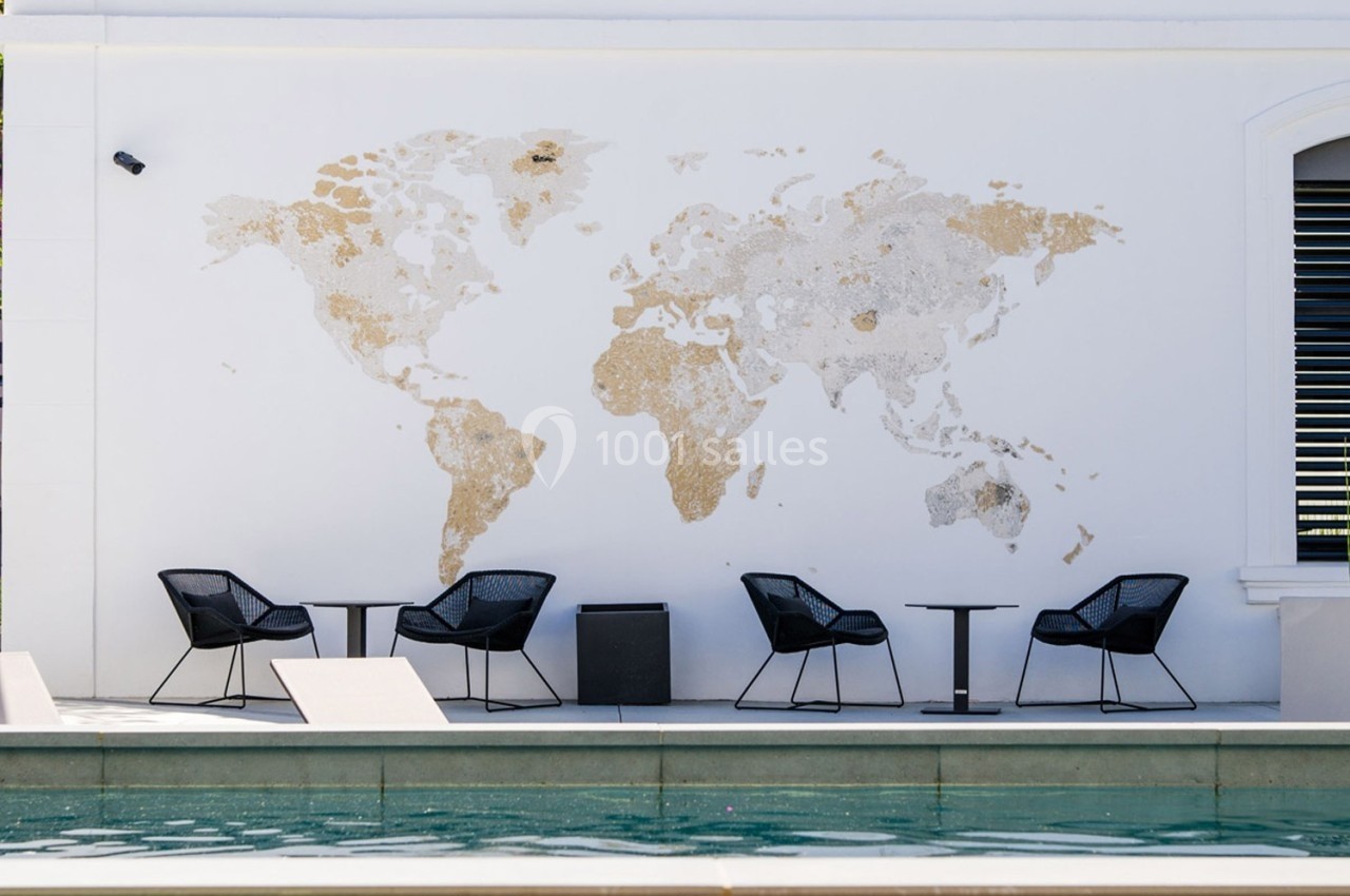 Mur blanc avec une carte du monde texturée, entouré de chaises noires et de petites tables près d'une piscine.