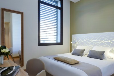 Chambre lumineuse avec lit double, table en bois, chaise beige, miroir et grande fenêtre avec stores.