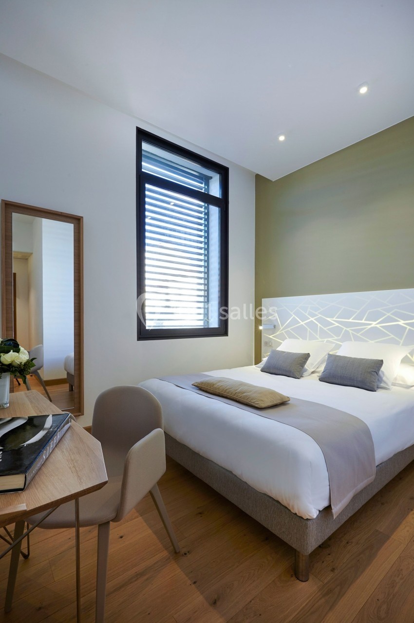 Chambre lumineuse avec lit double, table en bois, chaise beige, miroir et grande fenêtre avec stores.