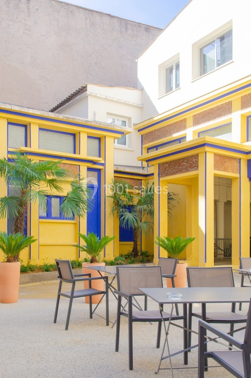 Cour intérieure lumineuse avec façades jaunes et bleues, palmiers en pots et mobilier de terrasse moderne.