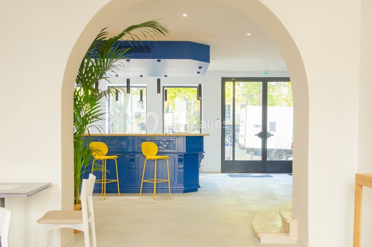 Intérieur lumineux avec un comptoir bleu, deux tabourets jaunes, une plante et une grande arche blanche.