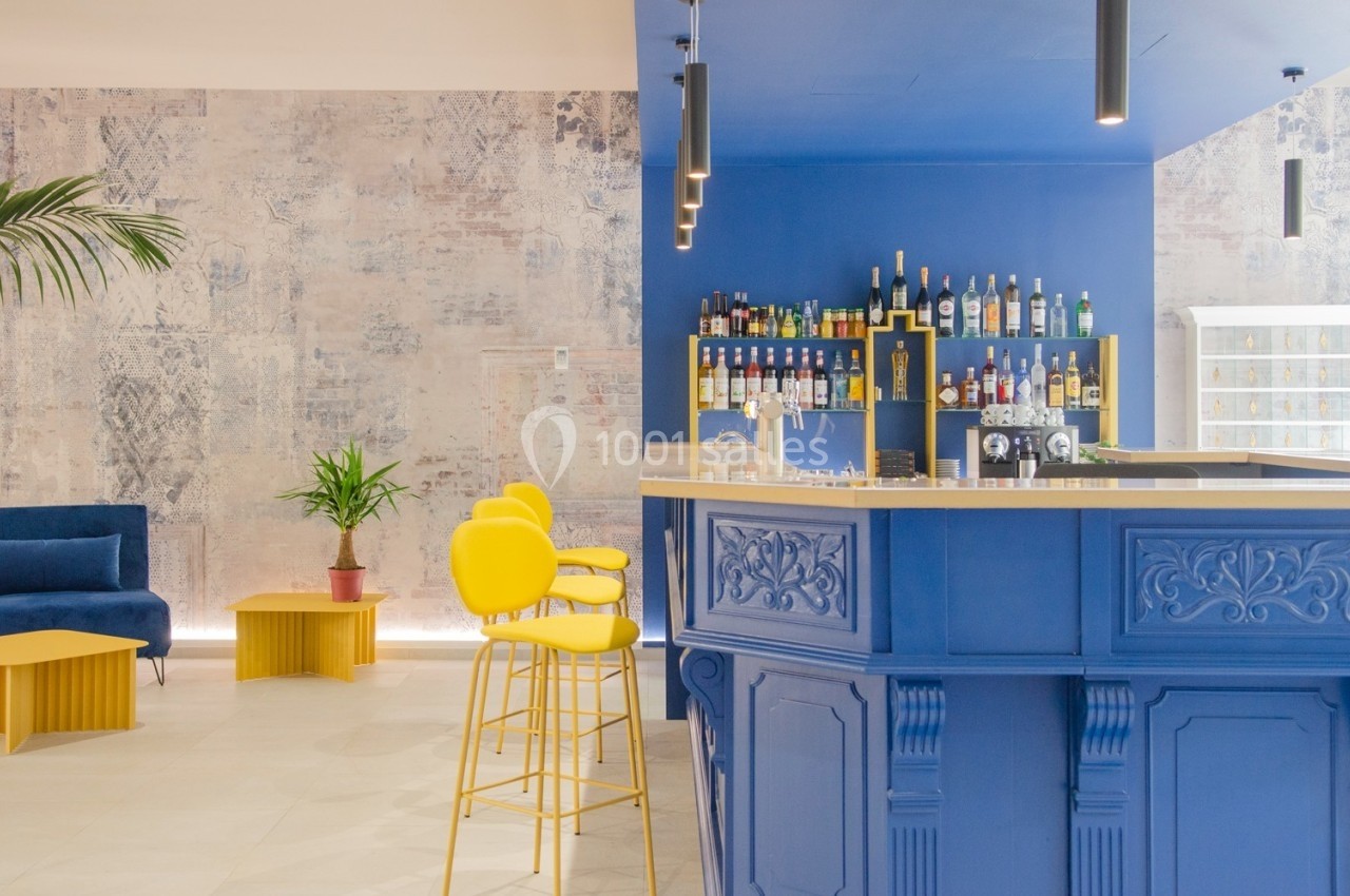 Bar moderne avec comptoir bleu, tabourets jaunes, étagères de bouteilles et mur texturé en arrière-plan.