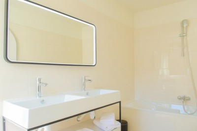 Salle de bain lumineuse avec un double lavabo, un grand miroir, une baignoire et des murs carrelés clairs.