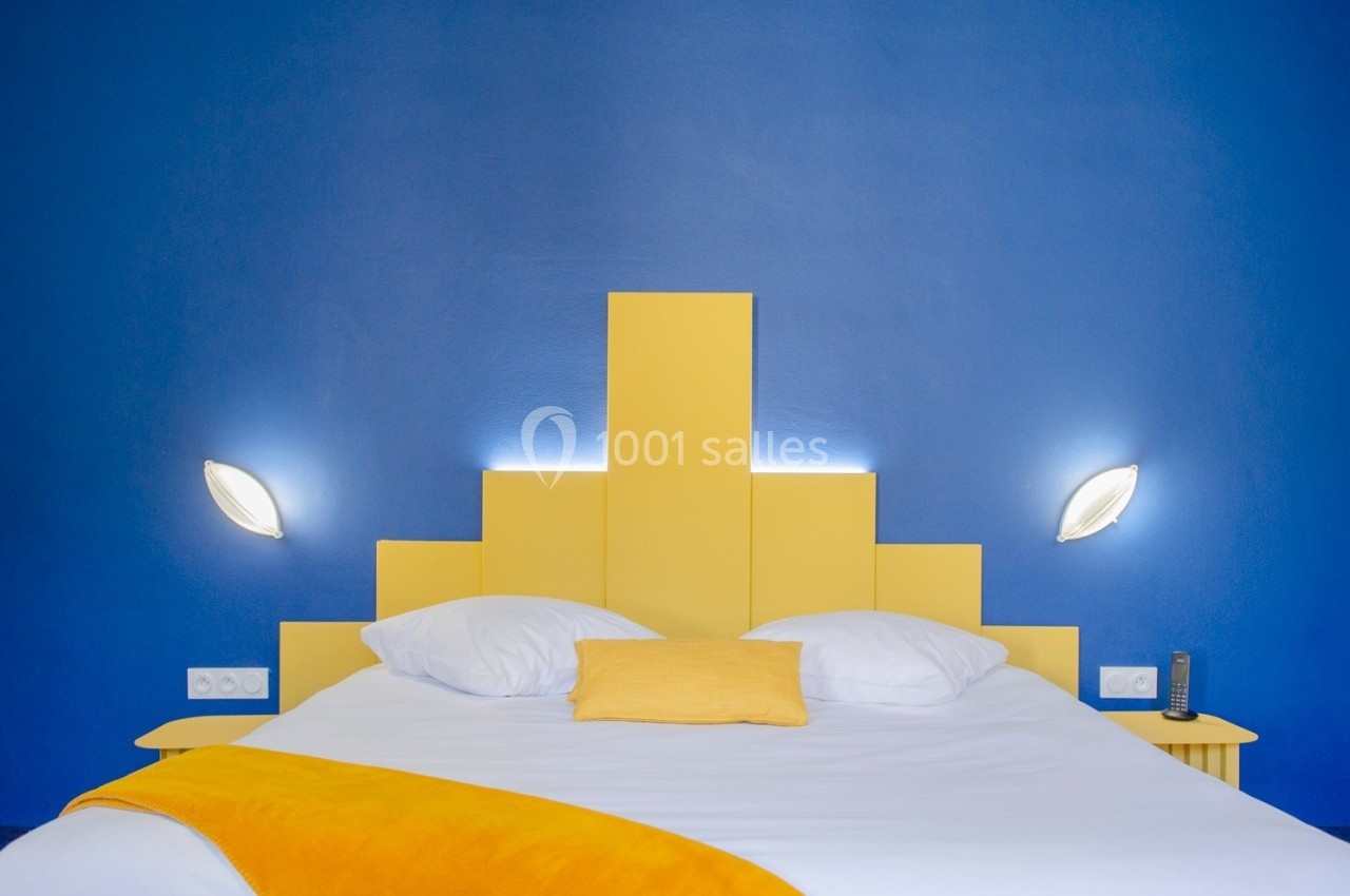 Lit double avec tête de lit jaune géométrique, draps blancs et couverture jaune, sur un fond de mur bleu vif.