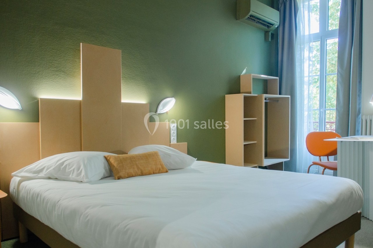 Chambre moderne avec lit double, mobilier en bois clair, mur vert, bureau orange et fenêtre donnant sur l'extérieur.