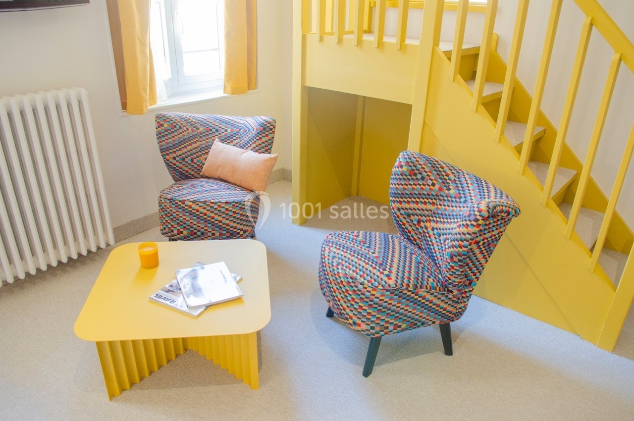 Deux fauteuils colorés, une table basse jaune avec des magazines et une tasse, près d'un escalier et d'une fenêtre.