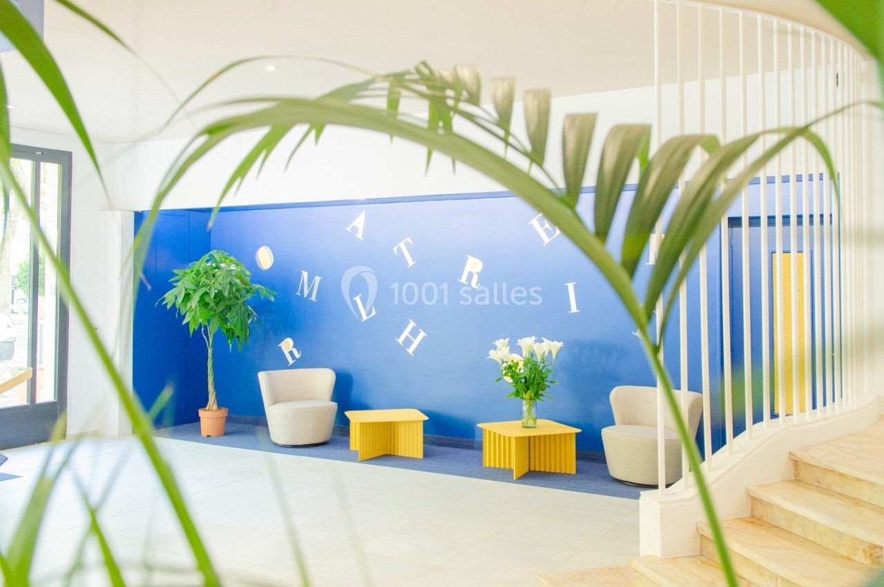 Espace d'accueil lumineux avec fauteuils, tables jaunes, plantes et mur bleu décoré de lettres blanches.