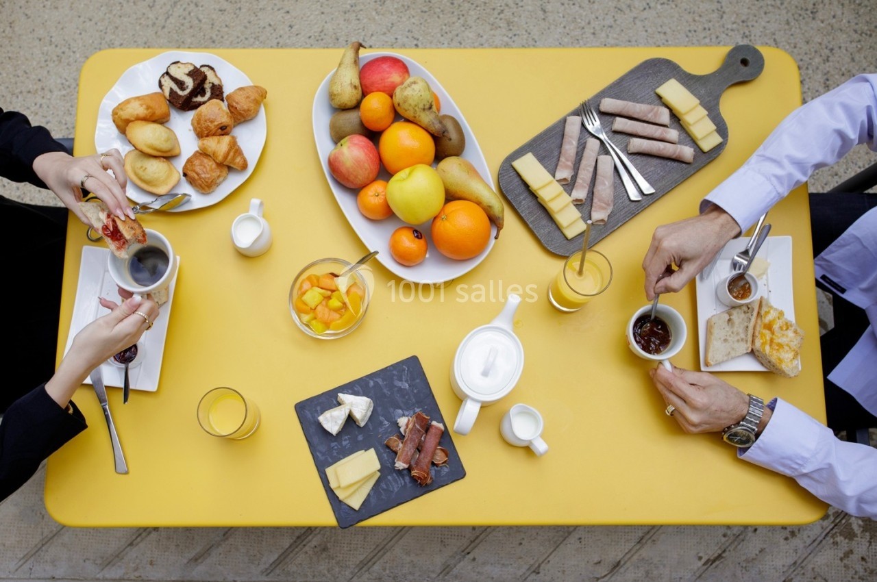 Petit-déjeuner avec fruits, viennoiseries, fromages, charcuterie et boissons, disposés sur une table jaune.