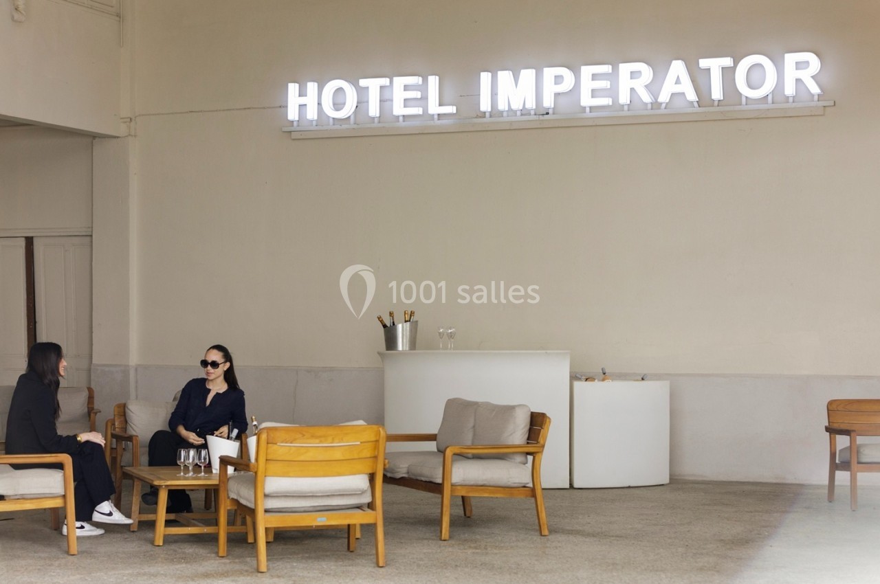 Deux femmes discutent assises sur des fauteuils en bois sous une enseigne lumineuse ’Hotel Imperator’.