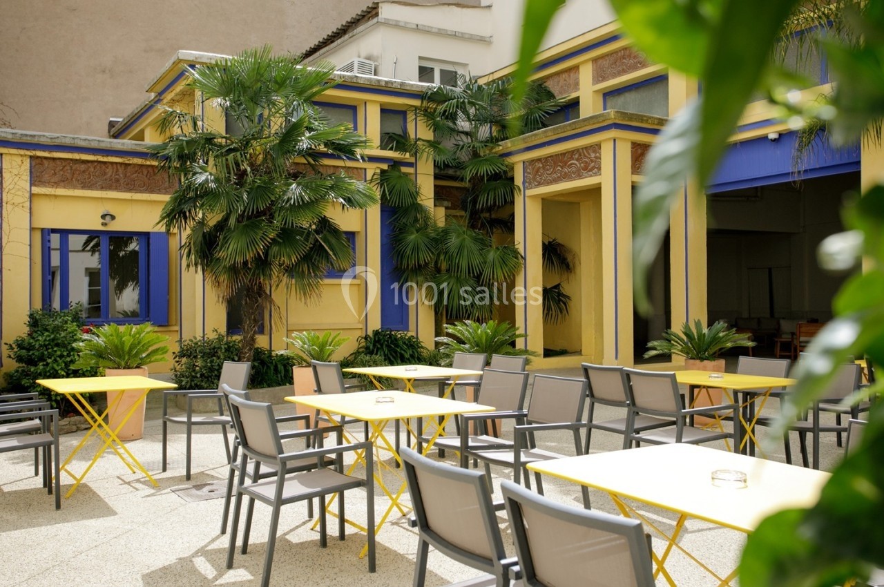Cour intérieure lumineuse avec tables et chaises jaunes et grises, entourée de plantes et d'un bâtiment coloré.