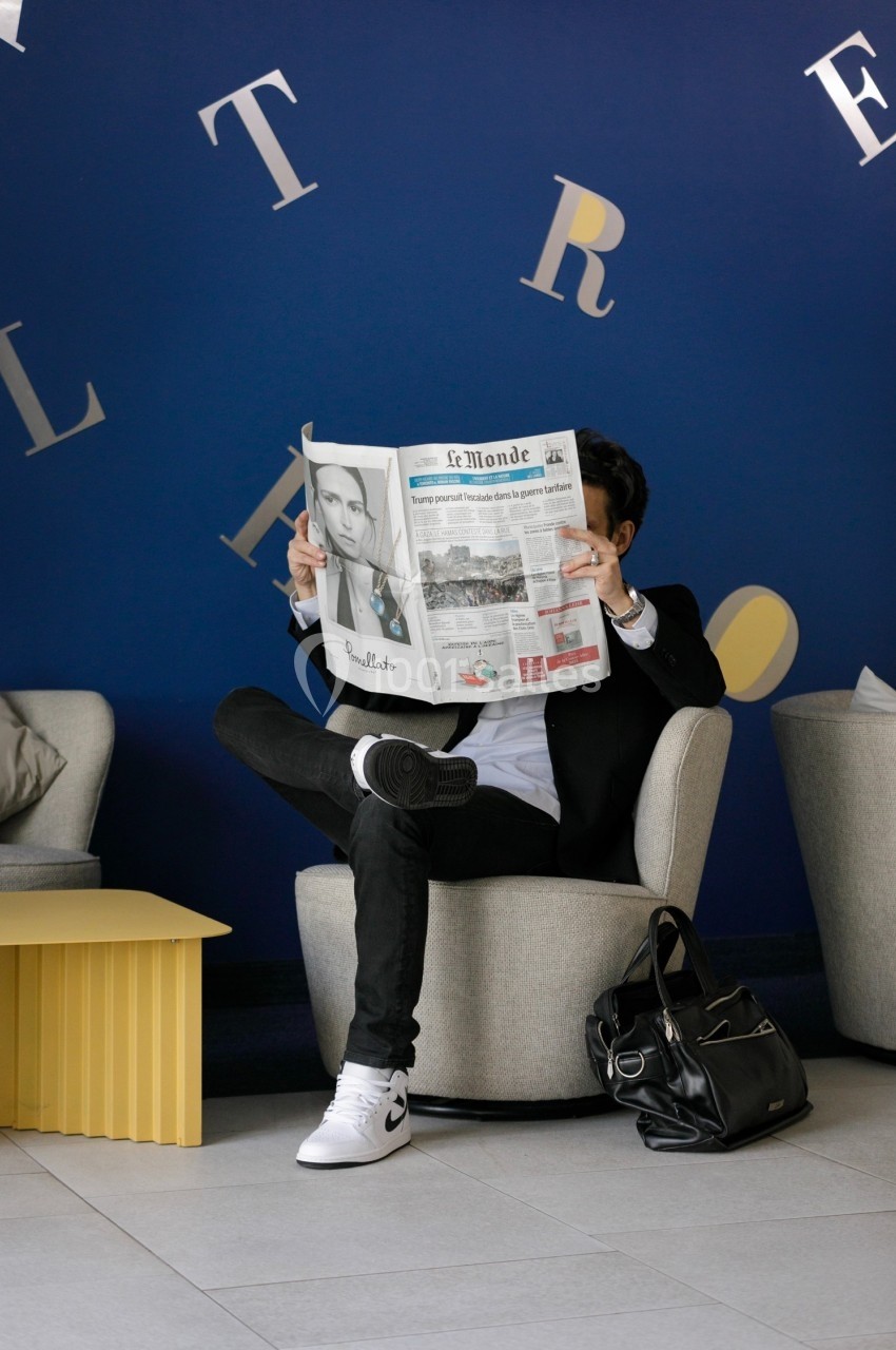 Un homme assis dans un fauteuil lit un journal dans un espace moderne avec un mur bleu en arrière-plan.