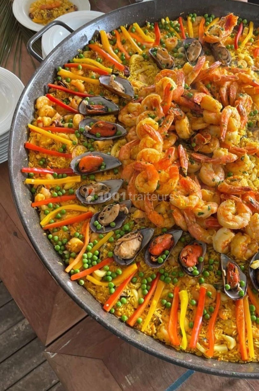 Grande poêle de paella garnie de crevettes, moules, légumes colorés et riz safrané, posée sur une table en bois.