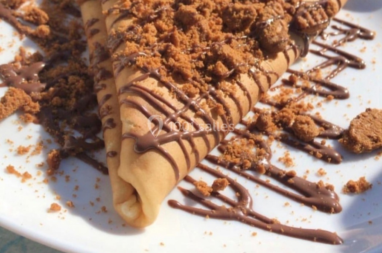 Crêpe pliée garnie de chocolat fondu et de miettes de biscuits, servie sur une assiette blanche.