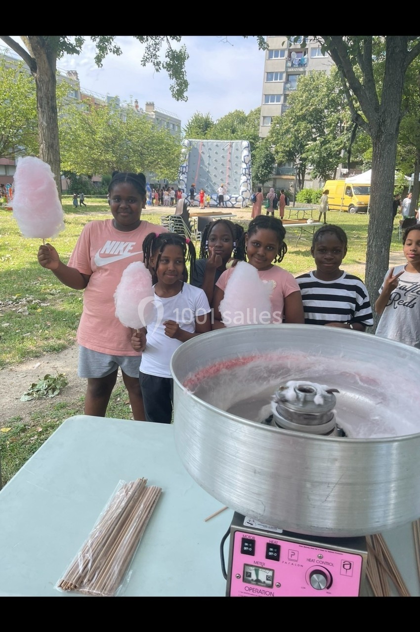 Des enfants souriants tiennent des barbes à papa dans un parc, près d'une machine à barbe à papa sur une table.