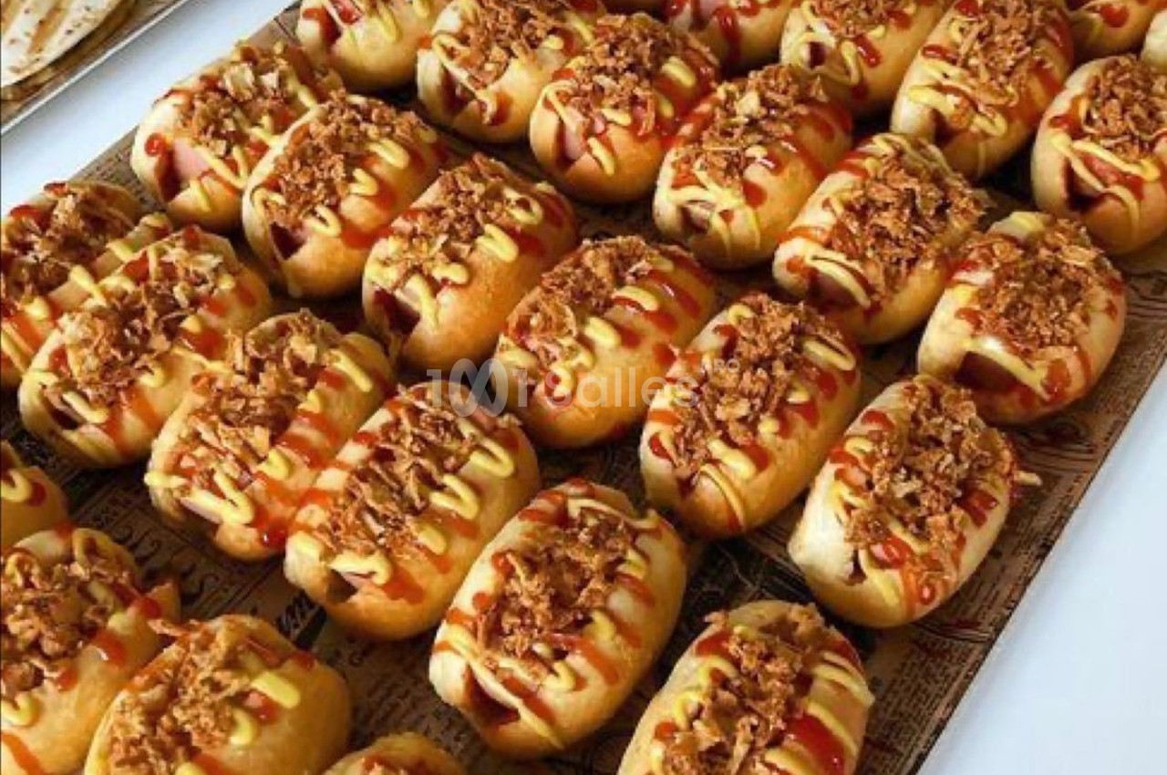 Rangées de petits pains garnis de sauce, moutarde et oignons frits, disposés sur un plateau.