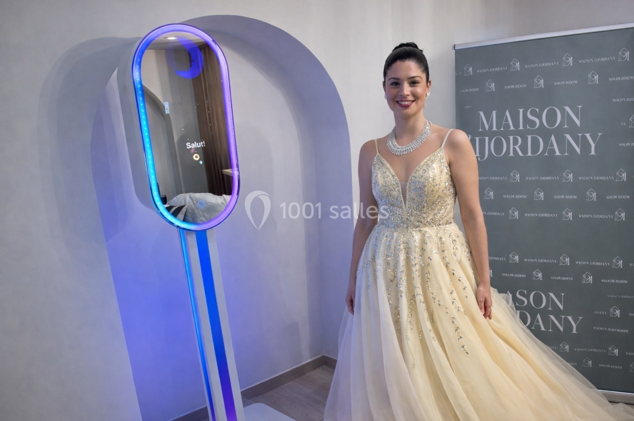 Femme en robe de soirée beige ornée, posant près d'un miroir interactif lumineux dans un intérieur élégant.