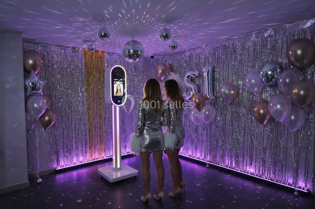 Deux femmes en robes à paillettes posent devant un photobooth lumineux dans une pièce décorée de ballons et guirlandes…