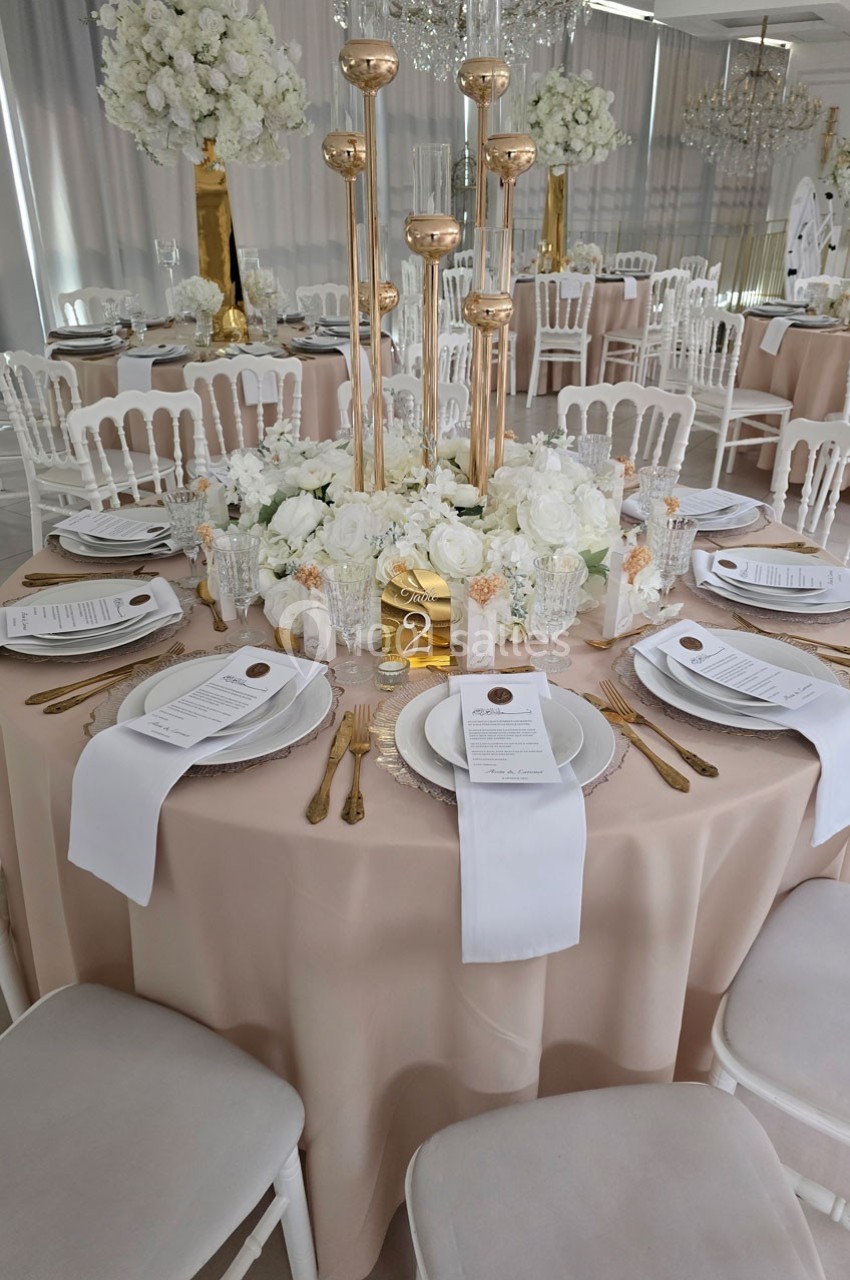 Table élégamment dressée avec nappes beige, vaisselle raffinée, bougies dorées et centre de table floral blanc.