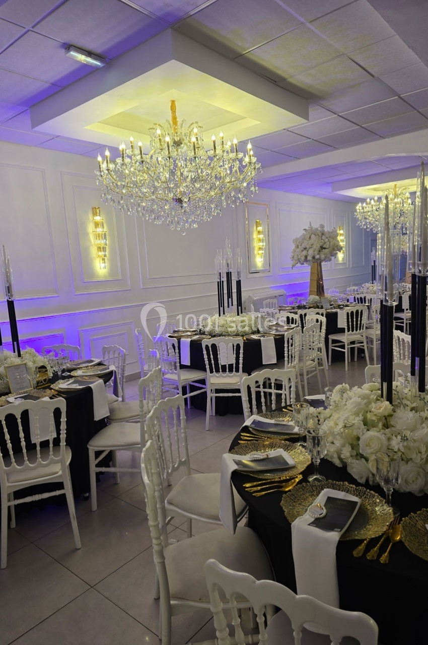 Salle de réception élégante avec tables décorées de nappes noires, chandeliers, fleurs blanches et lustres en cristal.