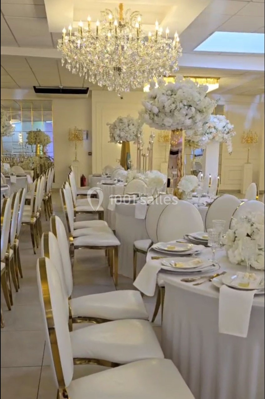 Salle de réception élégante avec tables décorées de fleurs blanches, chaises dorées et lustres en cristal.