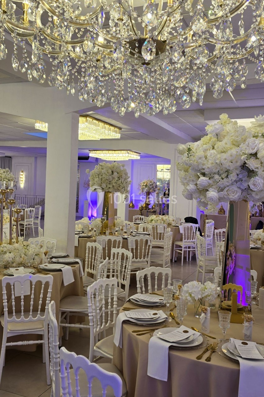 Salle de réception élégante avec tables décorées de nappes beiges, fleurs blanches et lustres en cristal suspendus.