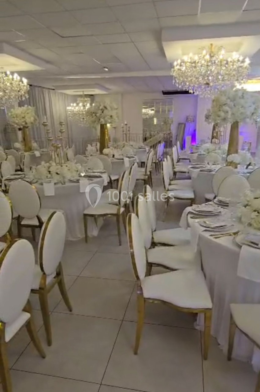 Salle de réception élégante avec tables rondes dressées, chaises blanches et décorations florales blanches.