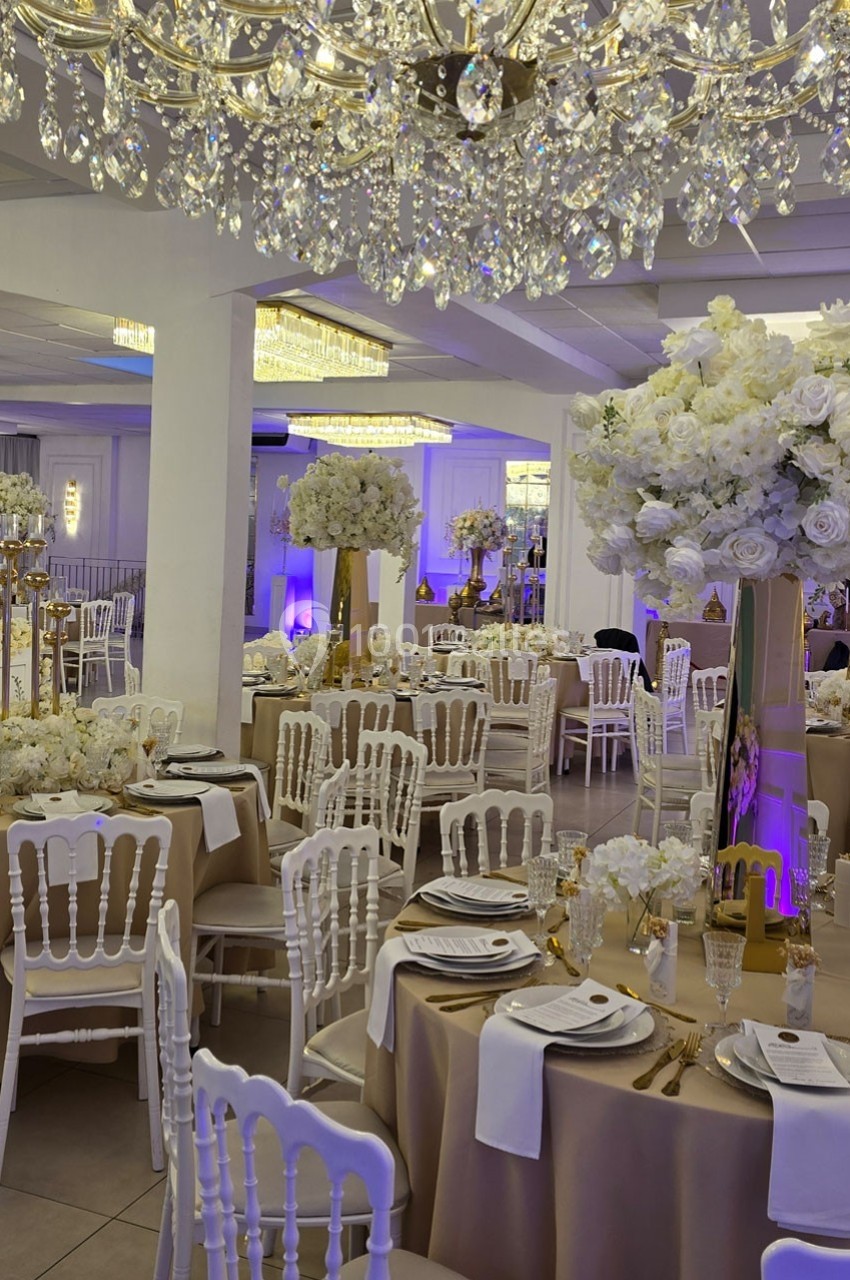 Salle de réception élégante décorée de chandeliers, fleurs blanches et tables dressées avec nappes beige et vaisselle…