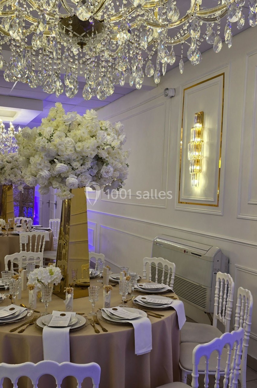 Salle de réception élégante avec tables décorées de nappes beige, centres de table fleuris et lustres en cristal.