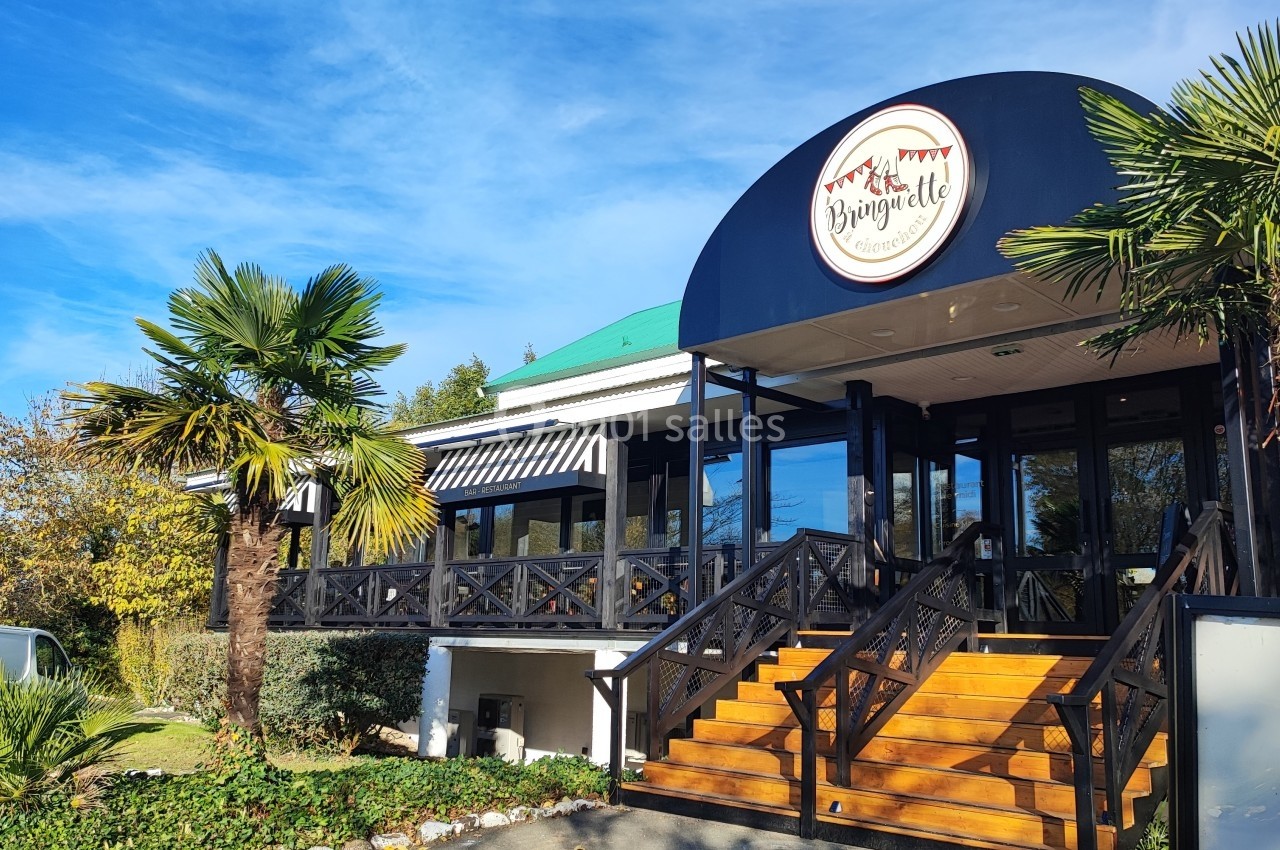 Entrée d'un restaurant avec terrasse en bois, palmiers et enseigne ronde sous un ciel bleu.