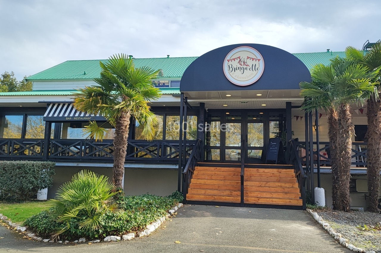 Façade d'un restaurant avec terrasse en bois, palmiers et toit vert, sous un ciel légèrement nuageux.