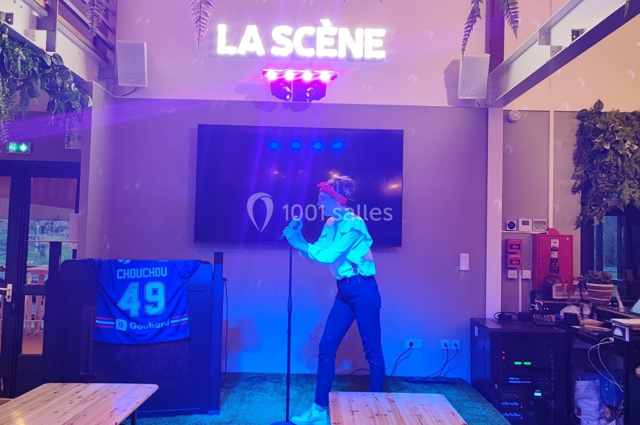 Une personne chante au micro sur une petite scène éclairée, avec un décor intérieur et un écran en arrière-plan.