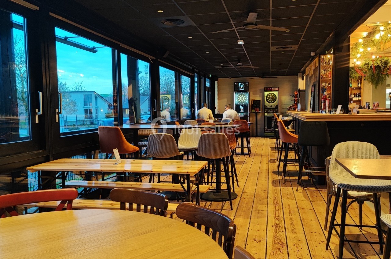 Salle de restaurant lumineuse avec grandes baies vitrées, tables en bois, chaises variées et un bar au fond.