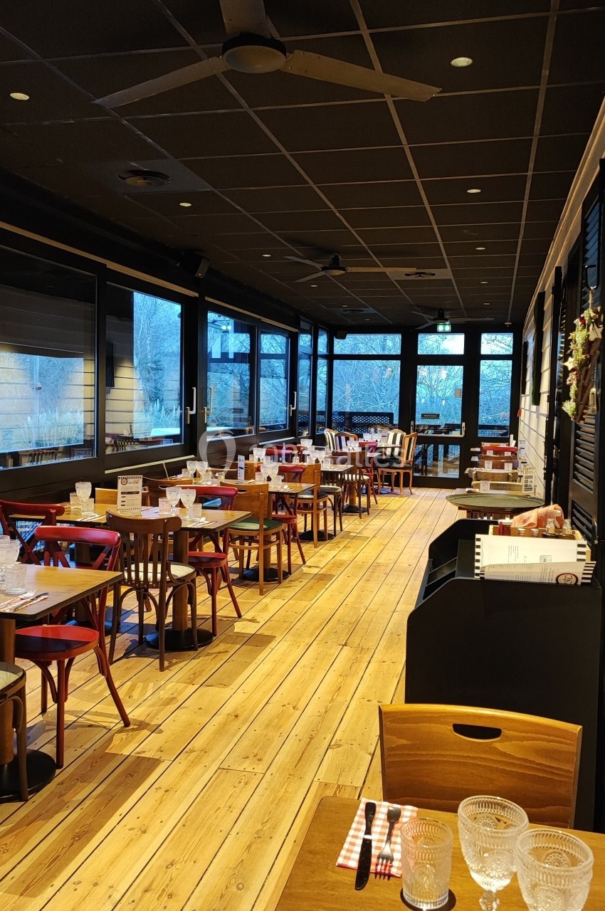 Salle de restaurant lumineuse avec tables en bois dressées, chaises rouges et grandes baies vitrées donnant sur l'extérieur.