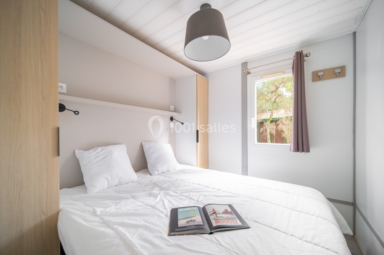 Chambre lumineuse avec lit double, draps blancs, étagère murale, fenêtre donnant sur un jardin et magazine ouvert sur le lit.