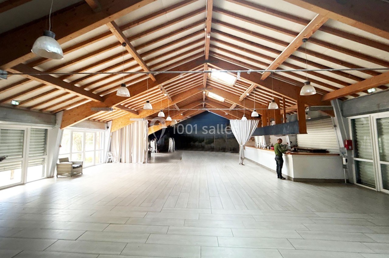 Salle spacieuse avec charpente en bois apparente, sol carrelé clair, bar à droite et grandes baies vitrées.