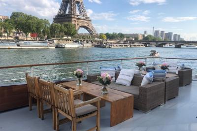 Terrasse d'un bateau avec des fauteuils, des plantes décoratives et une vue sur un pont et des arbres en arrière-plan.