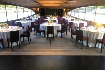 Terrasse d'un bateau avec des fauteuils, des plantes décoratives et une vue sur un pont et des arbres en arrière-plan.