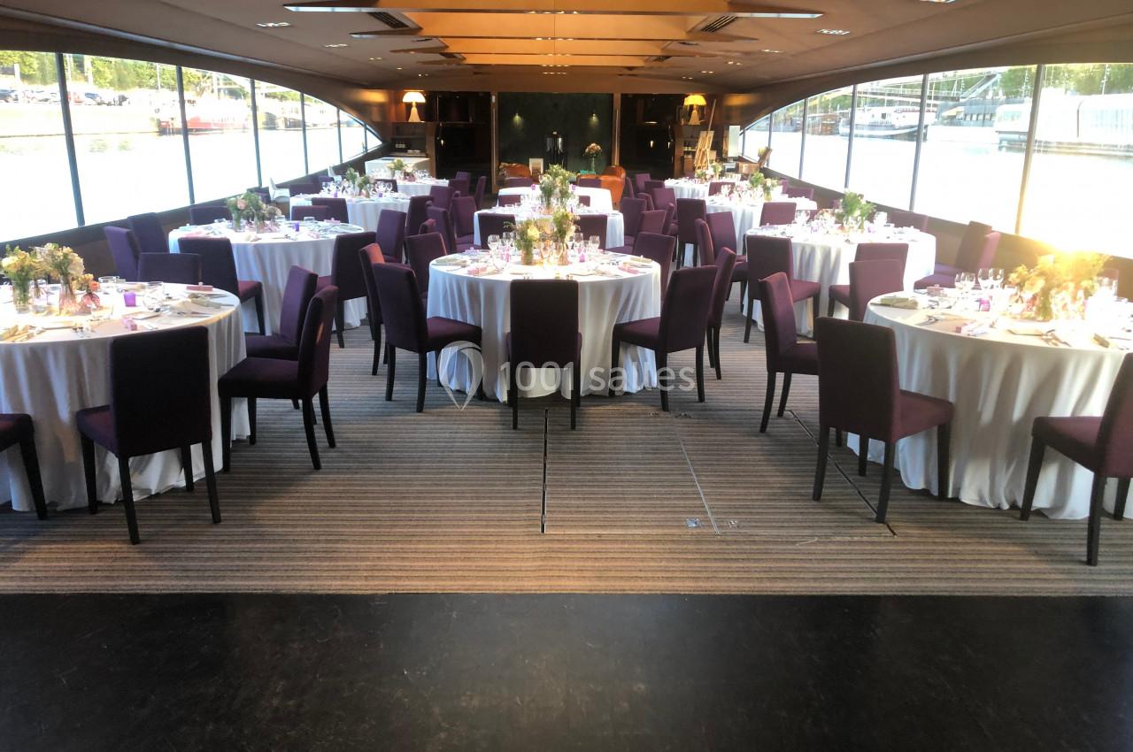Salle de réception élégante avec tables rondes dressées, nappes blanches et chaises violettes, éclairée par la lumière…