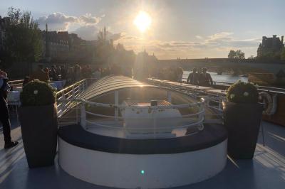 Terrasse d'un bateau avec des fauteuils, des plantes décoratives et une vue sur un pont et des arbres en arrière-plan.