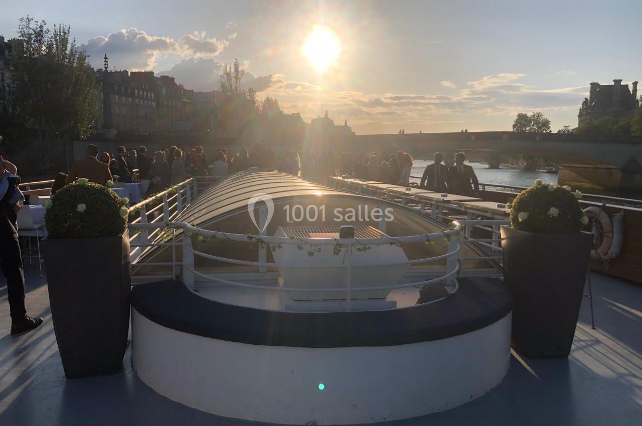 Pont d'un bateau au coucher du soleil, avec des passagers rassemblés et un pont visible à l'arrière-plan.