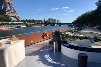 Terrasse d'un bateau avec des fauteuils, des plantes décoratives et une vue sur un pont et des arbres en arrière-plan.