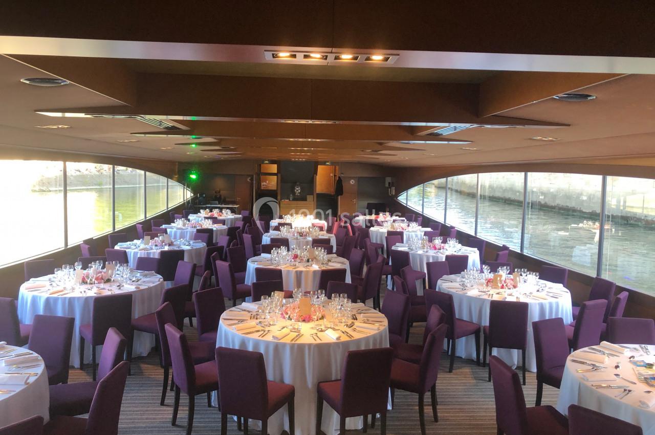 Salle de réception sur un bateau avec des tables rondes dressées, nappes blanches et chaises violettes, vue sur l'eau.