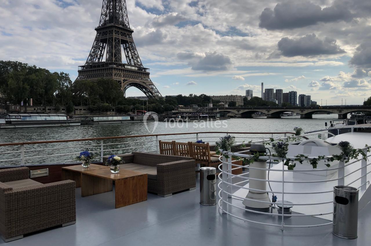 Vue d'un pont de bateau aménagé avec des sièges et des décorations florales, face à la tour Eiffel et la Seine.